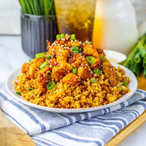 A plate of fried rice topped with pieces of glazed chicken, garnished with chopped green onions and sesame seeds, sits on a striped cloth with a wooden spoon beside it.