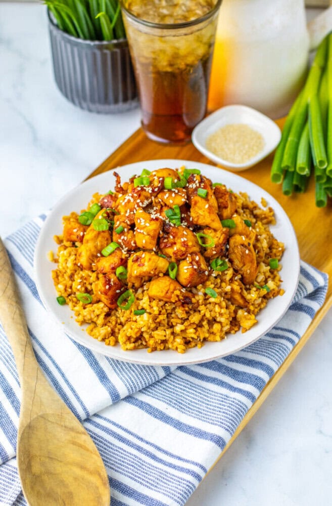 A plate of fried rice topped with sesame chicken and green onions sits on a striped towel, with a wooden spoon, green onions, soda, and a small dish of sesame seeds nearby.