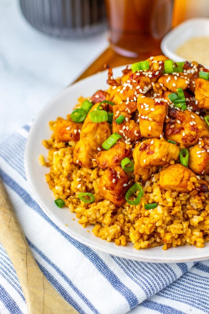 A white plate of diced chicken in sauce served over rice, topped with sesame seeds and chopped green onions, placed on a striped napkin.