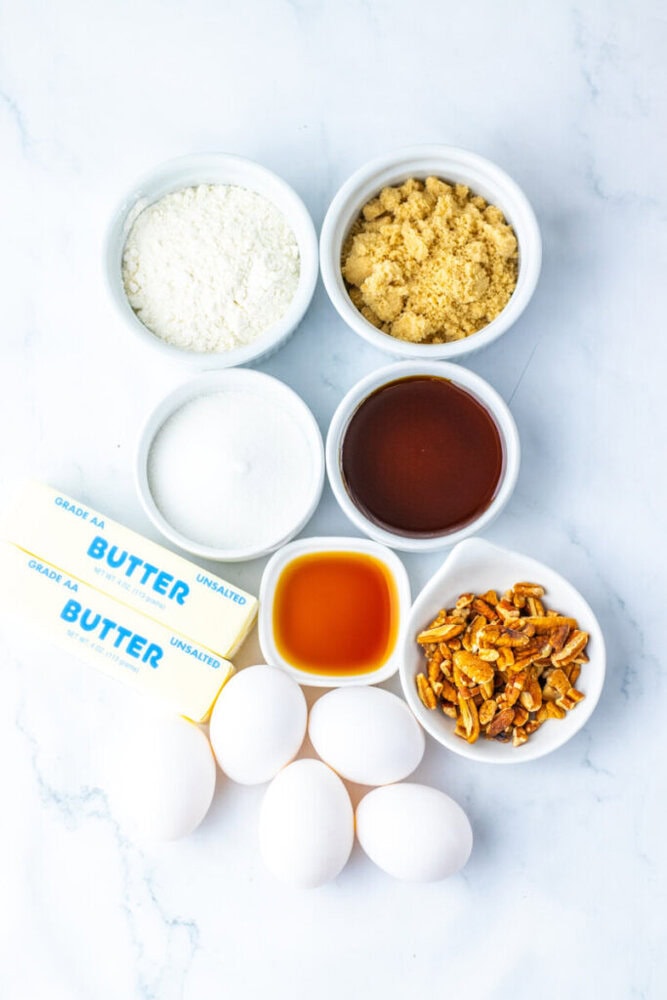 Top-down view of baking ingredients: flour, brown sugar, white sugar, maple syrup, vanilla extract, butter sticks, eggs, and chopped pecans arranged on a white surface.