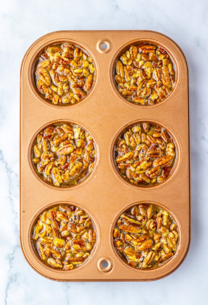 A gold muffin tin on a marble surface containing six unbaked pecan pie fillings.