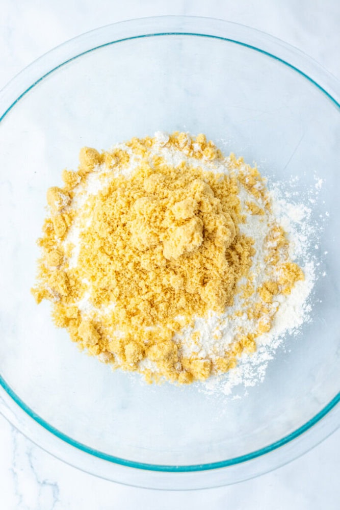 A clear glass bowl containing a mixture of flour and brown sugar on a white marble surface.