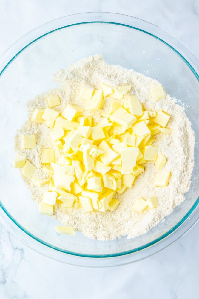 A glass bowl containing flour and small cubes of butter on a light-colored surface.