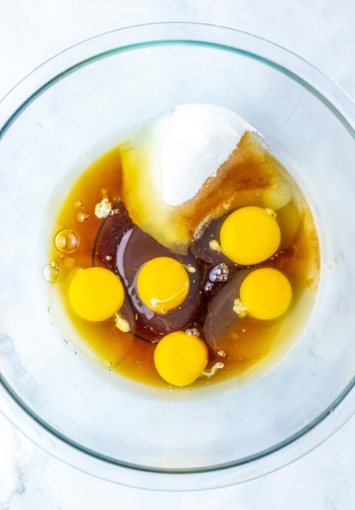 A glass bowl containing six raw eggs, sugar, vanilla extract, and another liquid ingredient, viewed from above on a white surface.