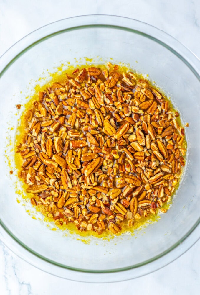 A glass bowl filled with chopped pecans mixed in a yellow liquid, likely melted butter or oil, on a white surface.