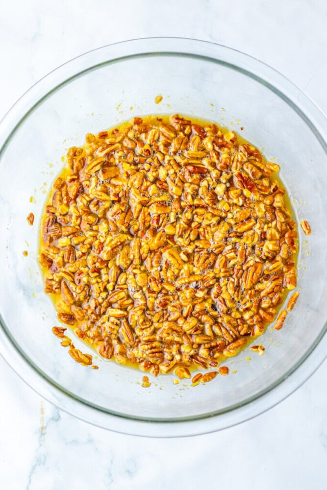A glass bowl filled with chopped pecans coated in a yellow-orange mixture, likely butter and sugar, on a white surface.