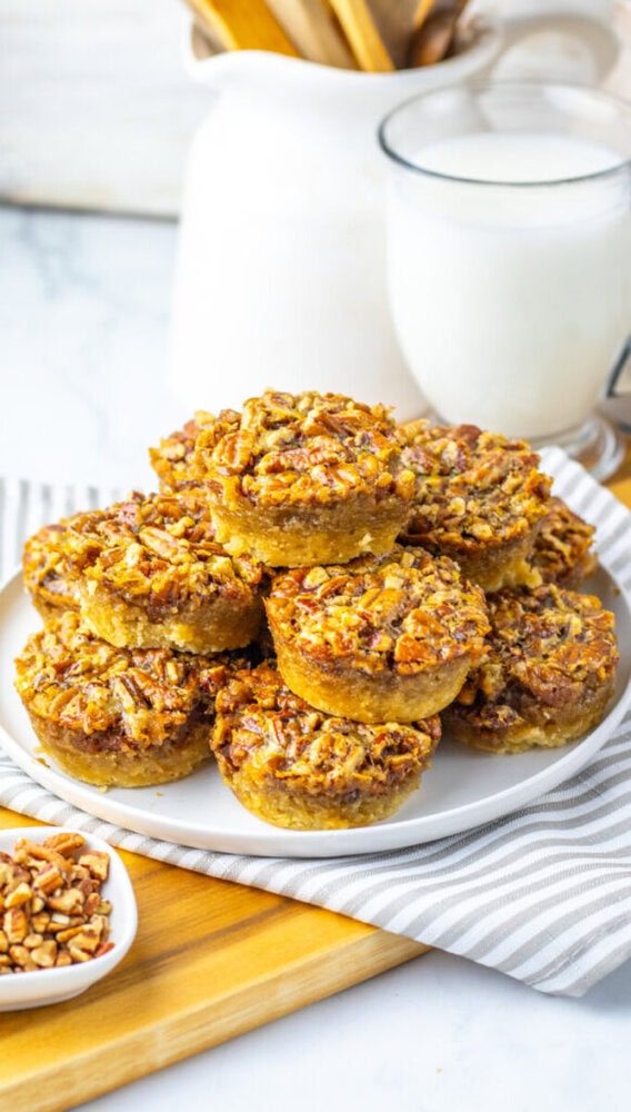 A plate stacked with mini pecan pies sits on a striped cloth, with a small bowl of chopped pecans and a glass of milk in the background.