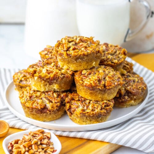 A plate of pecan pie muffins stacked on a white plate, with a small bowl of chopped pecans, wooden measuring spoons, and a glass of milk in the background.