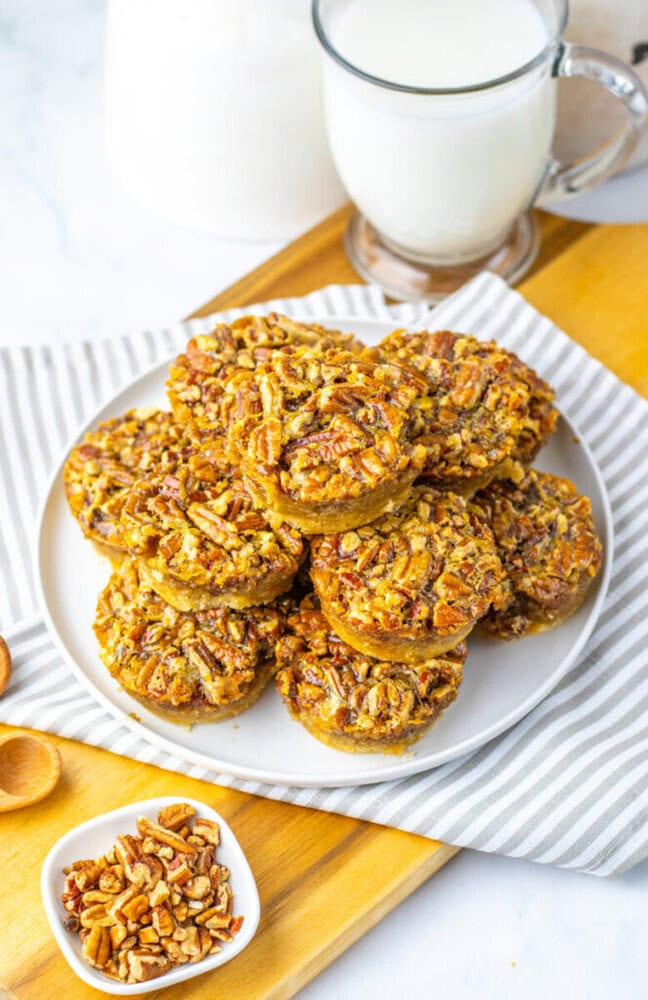 A plate of mini pecan pies is arranged on a white plate, with a glass of milk and a small bowl of chopped pecans nearby.