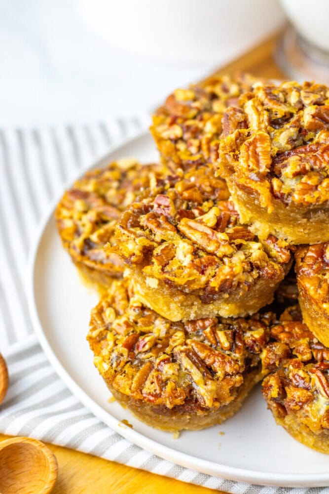 A plate of mini pecan pies stacked on top of each other, showing a nutty filling and golden crust, set on a white and striped background.