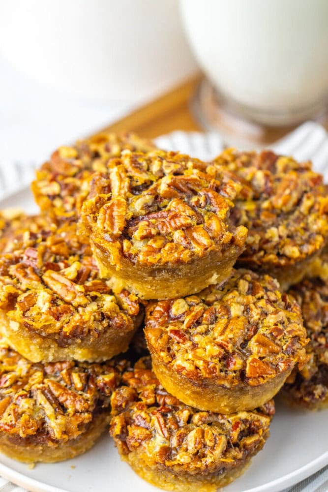 A plate of mini pecan pies is stacked on a white dish, with glasses of milk in the background.
