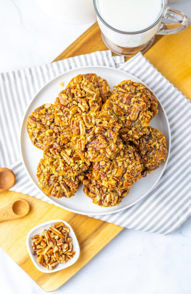 A plate of pecan cookies sits on a striped cloth, with a small bowl of chopped pecans and a glass of milk nearby on a wooden board.