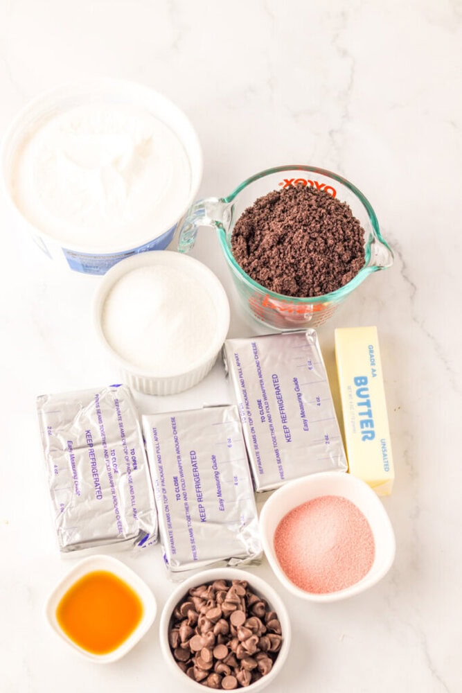 An assortment of cheesecake ingredients on a marble surface, including cream cheese, sugar, butter, whipped topping, chocolate chips, crushed cookies, pink sugar, and vanilla extract.
