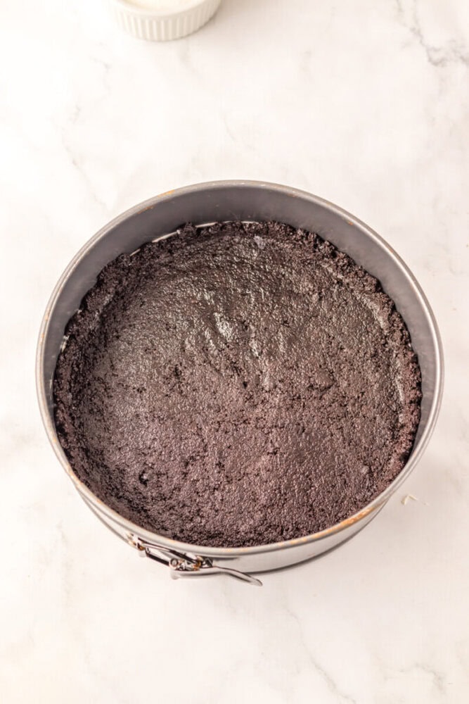 A round springform pan filled with a pressed chocolate cookie crust on a white marble surface.