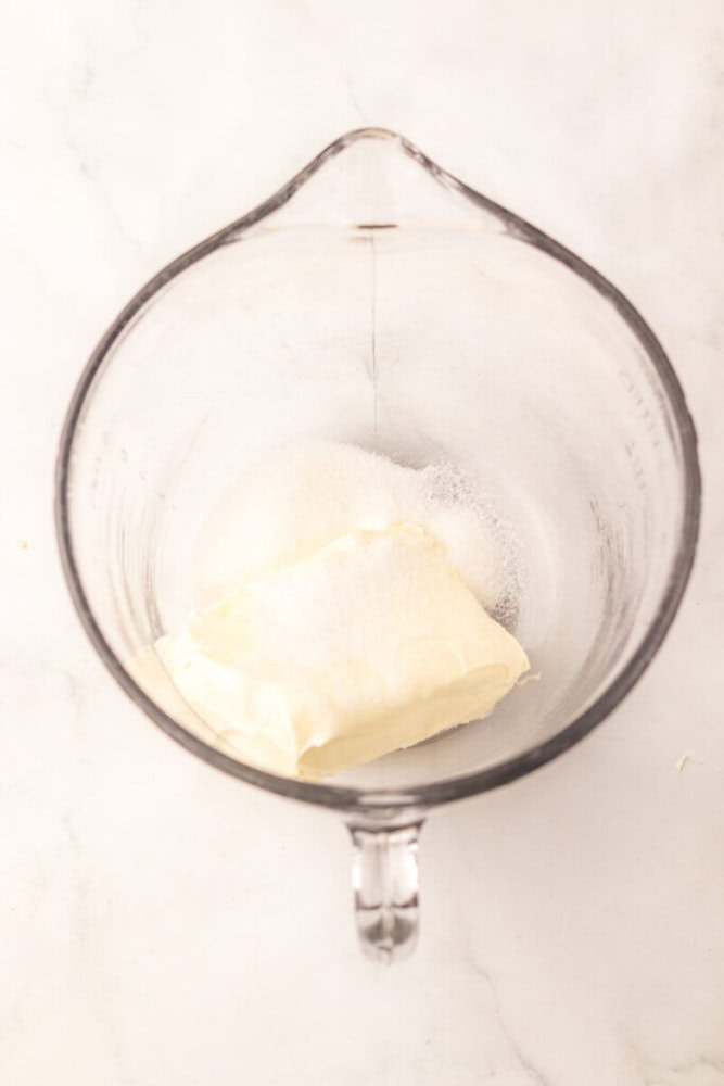 A glass mixing bowl containing a block of cream cheese and a pile of granulated sugar on a white surface.