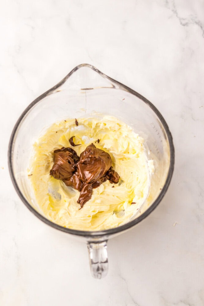 A glass mixing bowl containing whipped butter and dollops of chocolate hazelnut spread on a marble surface.