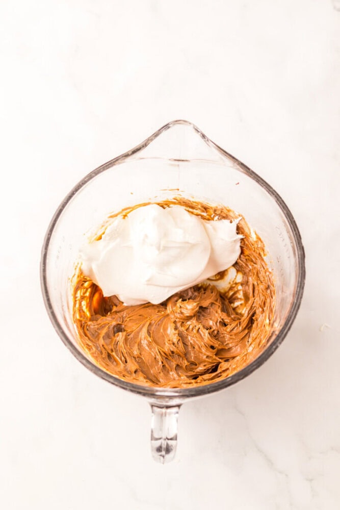 A glass mixing bowl containing whipped cream on top of a layer of blended brown mixture, set on a white surface.