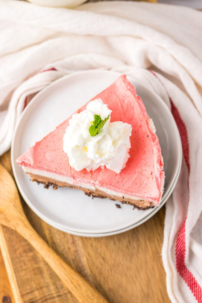 A slice of pink cheesecake with a chocolate crust, topped with whipped cream and a mint leaf, on a white plate.