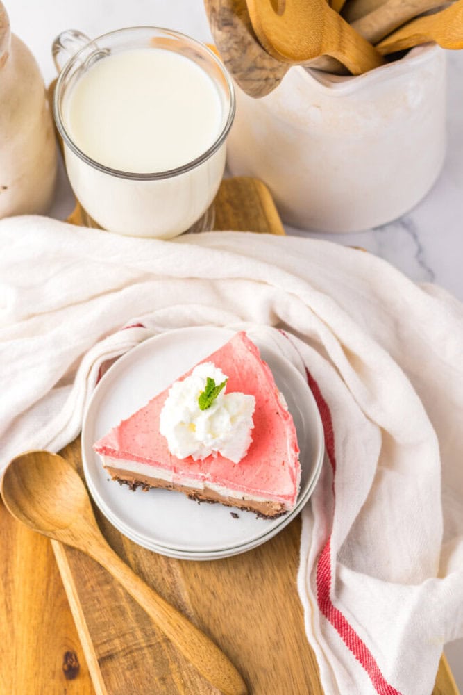 A slice of pink cheesecake with whipped cream and a mint leaf, served on a plate with a wooden spoon, next to a glass of milk and kitchen utensils.