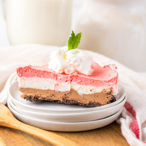 A slice of layered chocolate, vanilla, and strawberry dessert topped with whipped cream and a mint leaf, served on a white plate with a wooden spoon beside it.