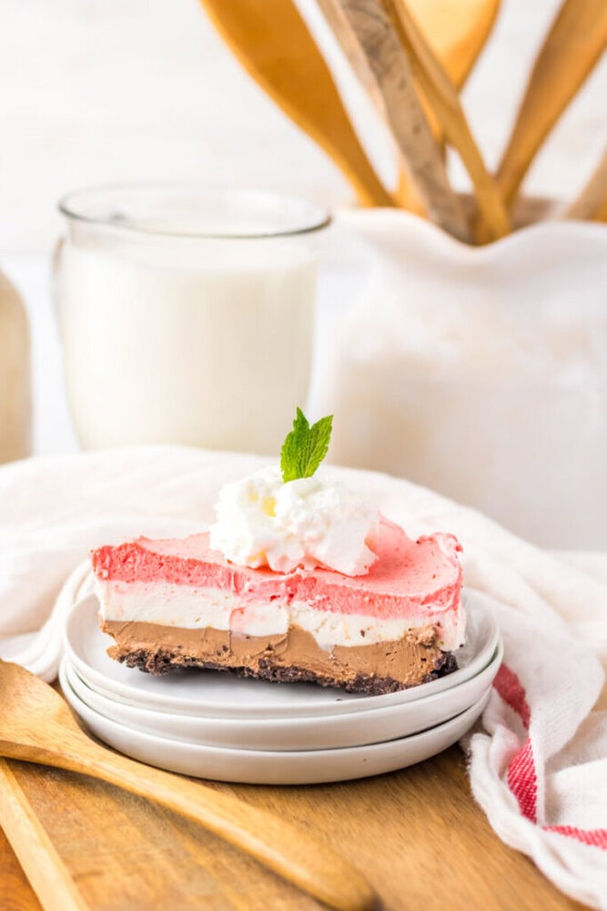 A slice of layered chocolate and strawberry dessert topped with whipped cream and a mint leaf sits on stacked plates, with a glass of milk and utensils in the background.