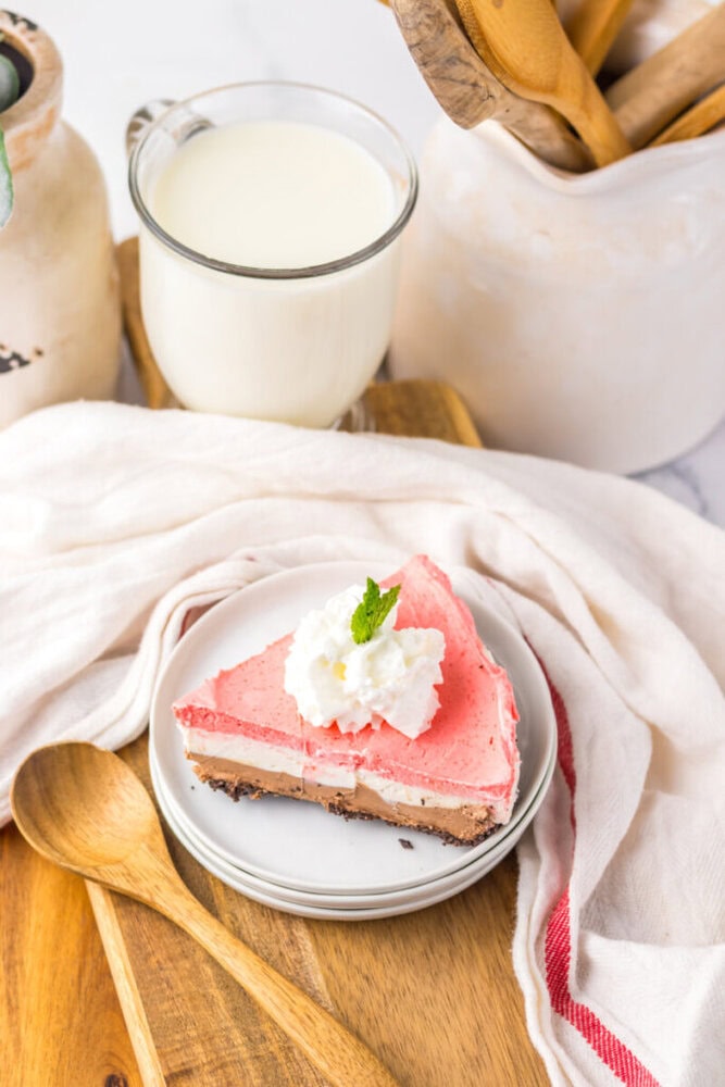 A slice of pink cheesecake with whipped cream and a mint leaf on a plate, next to a glass of milk and wooden spoons on a wooden surface.