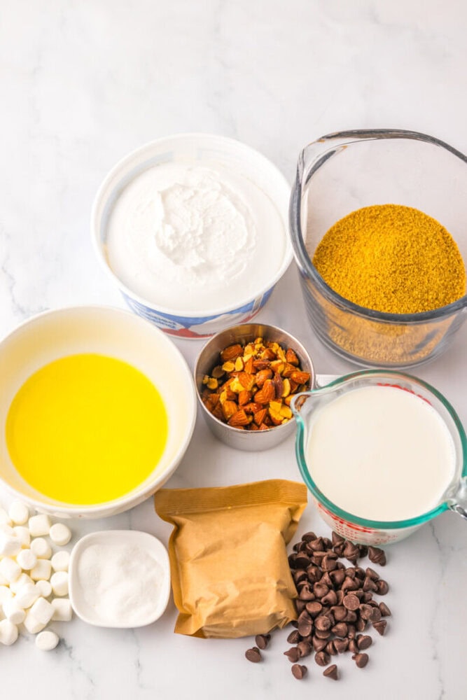 Various baking ingredients including melted butter, yogurt, graham cracker crumbs, chopped nuts, milk, mini marshmallows, baking powder, and chocolate chips arranged on a marble surface.