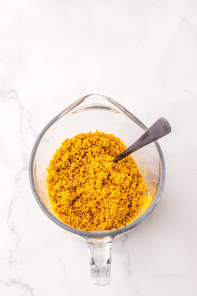 A glass measuring cup filled with a yellow, crumbly mixture and a metal spoon on a white marble surface.