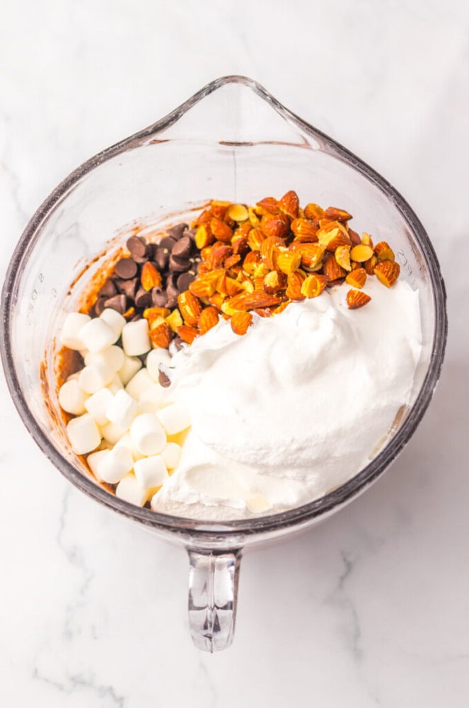 A glass mixing bowl containing marshmallows, chocolate chips, chopped almonds, and a mound of white powder on a marble surface.
