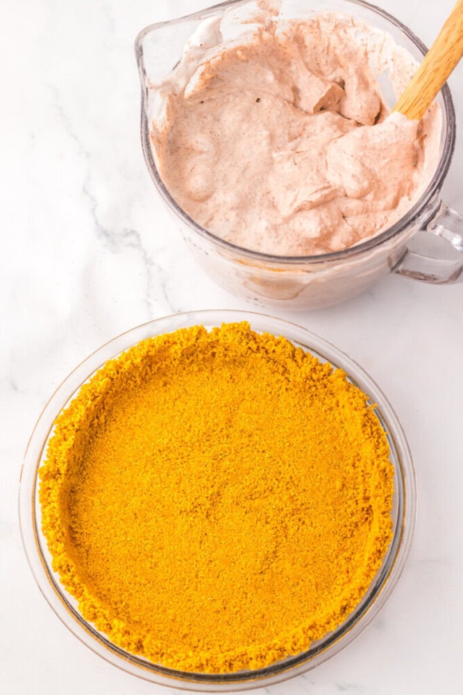 A glass bowl of creamy, light brown pie filling sits next to a pie dish with a golden graham cracker crust on a white surface.