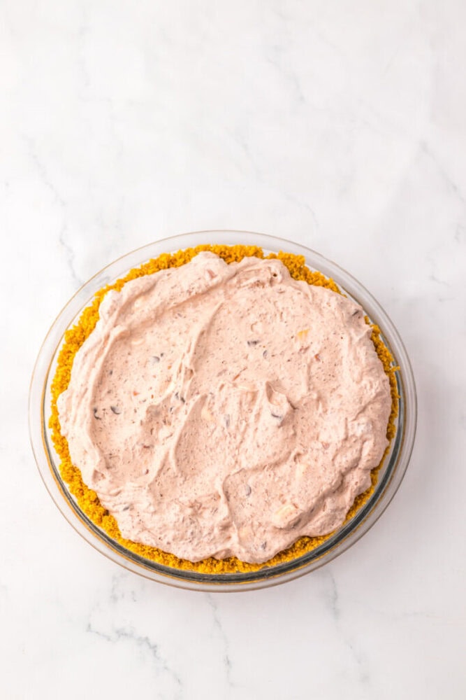 A pie with a light brown, creamy filling in a graham cracker crust, shown from above on a white marble surface.
