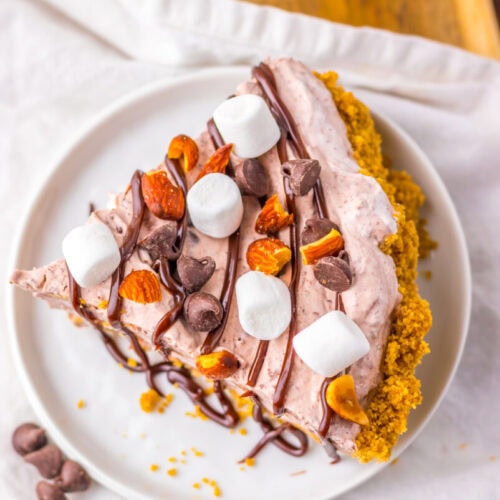 A slice of pie with a crumb crust, chocolate drizzle, mini marshmallows, chocolate chips, and almonds on a white plate.