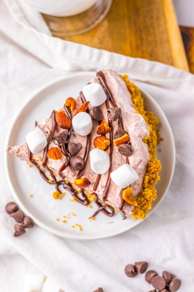 A slice of chocolate cream pie with graham cracker crust, topped with mini marshmallows, chocolate chips, chopped nuts, and a drizzle of chocolate sauce on a white plate.