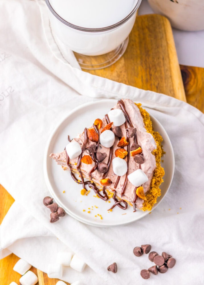 A slice of chocolate cream pie topped with marshmallows, chopped nuts, and chocolate drizzle on a white plate, with chocolate chips and marshmallows scattered nearby.