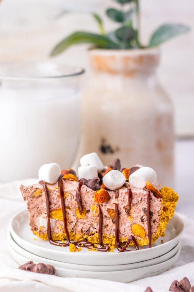A slice of chocolate and nut pie topped with mini marshmallows and chocolate drizzle, served on stacked white plates with a glass of milk and a vase in the background.
