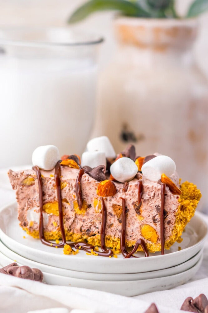 A slice of chocolate and marshmallow pie with a crumb crust, topped with mini marshmallows, nuts, chocolate chips, and a drizzle of chocolate sauce on a white plate.