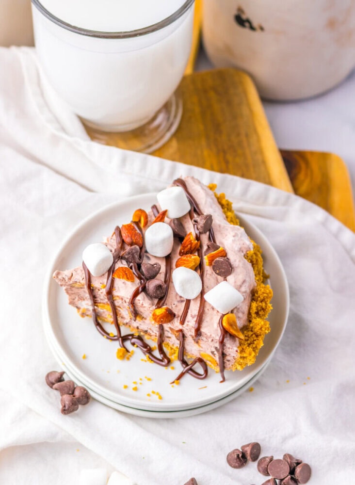 A slice of chocolate cream pie with a graham cracker crust, topped with mini marshmallows, nuts, chocolate chips, and chocolate drizzle, on a white plate next to a glass of milk.