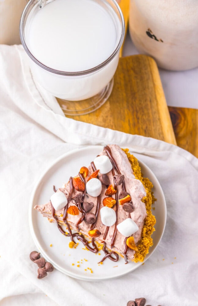 A slice of chocolate pie topped with mini marshmallows, chopped nuts, and chocolate drizzle on a white plate, next to a glass of milk.