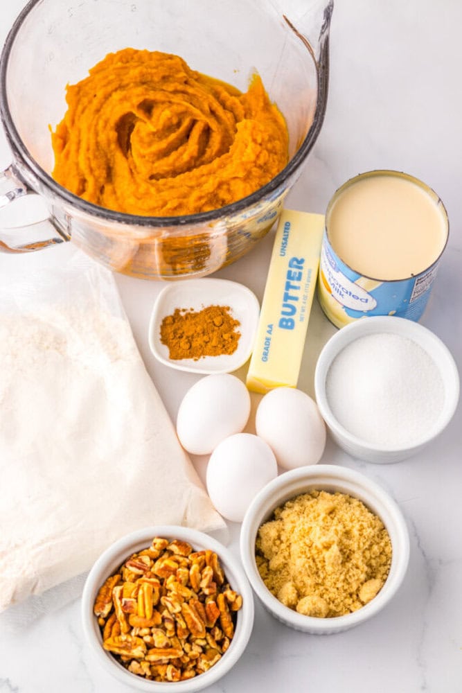 Ingredients for baking, including canned pumpkin, butter, sweetened condensed milk, flour, granulated sugar, brown sugar, eggs, chopped pecans, and spices, arranged on a counter.