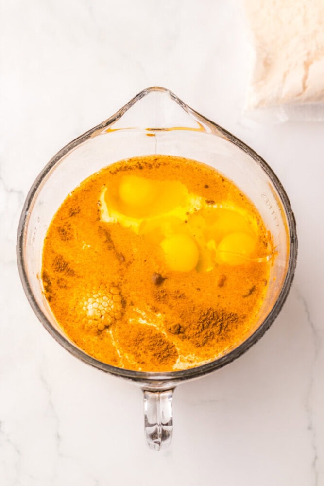 A glass measuring cup containing eggs, spices, and liquid ingredients on a white marble surface.