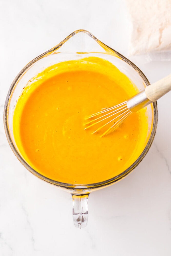A glass mixing bowl filled with yellow batter and a metal whisk resting inside on a white surface.
