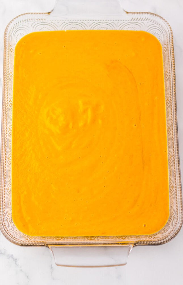 A glass baking dish filled with a smooth, bright orange batter or mixture, placed on a white marble surface.