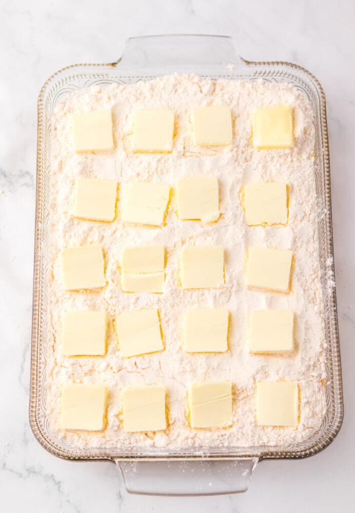 A glass baking dish filled with flour topped with evenly spaced square slices of butter on a white marble surface.