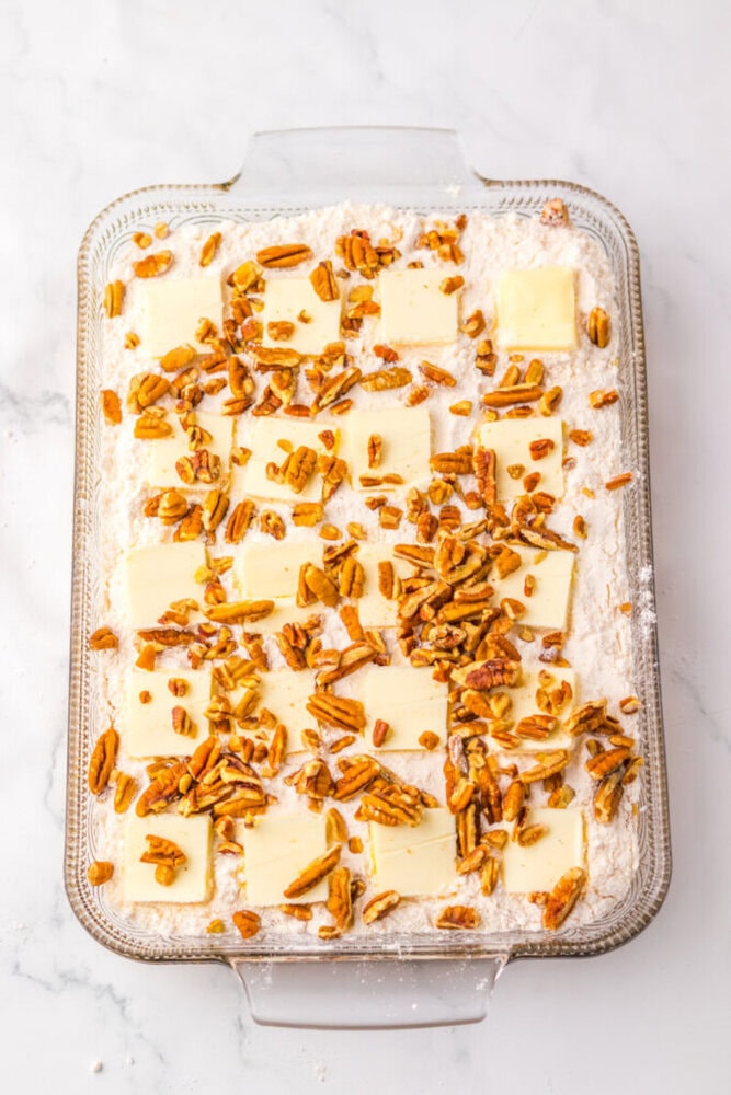 Glass baking dish with flour, butter slices, and chopped pecans arranged on top, set on a white marble surface.