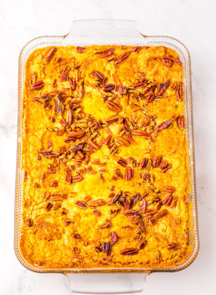 Rectangular glass baking dish filled with a golden-brown baked casserole topped with pecan pieces, placed on a white surface.