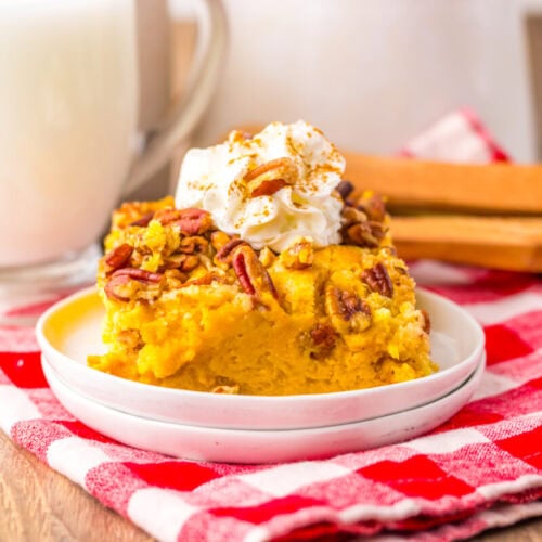 A slice of pumpkin dessert topped with pecans and whipped cream on a white plate, with a glass of milk in the background on a red and white checkered cloth.