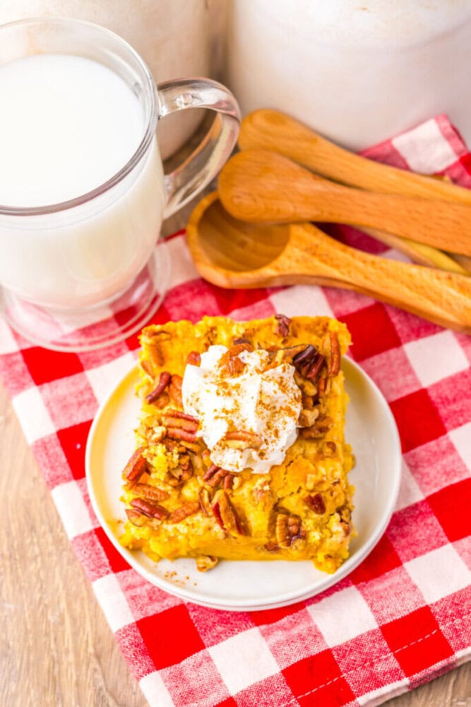 A slice of pumpkin dump cake topped with whipped cream and pecans on a white plate, next to a glass of milk and wooden spoons on a red and white checkered cloth.