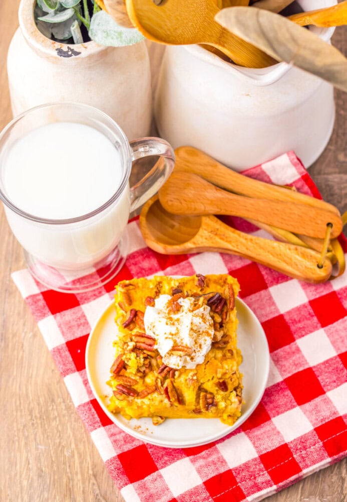 A slice of pumpkin dessert topped with whipped cream and pecans on a white plate, next to a glass of milk and wooden utensils on a red and white checkered napkin.