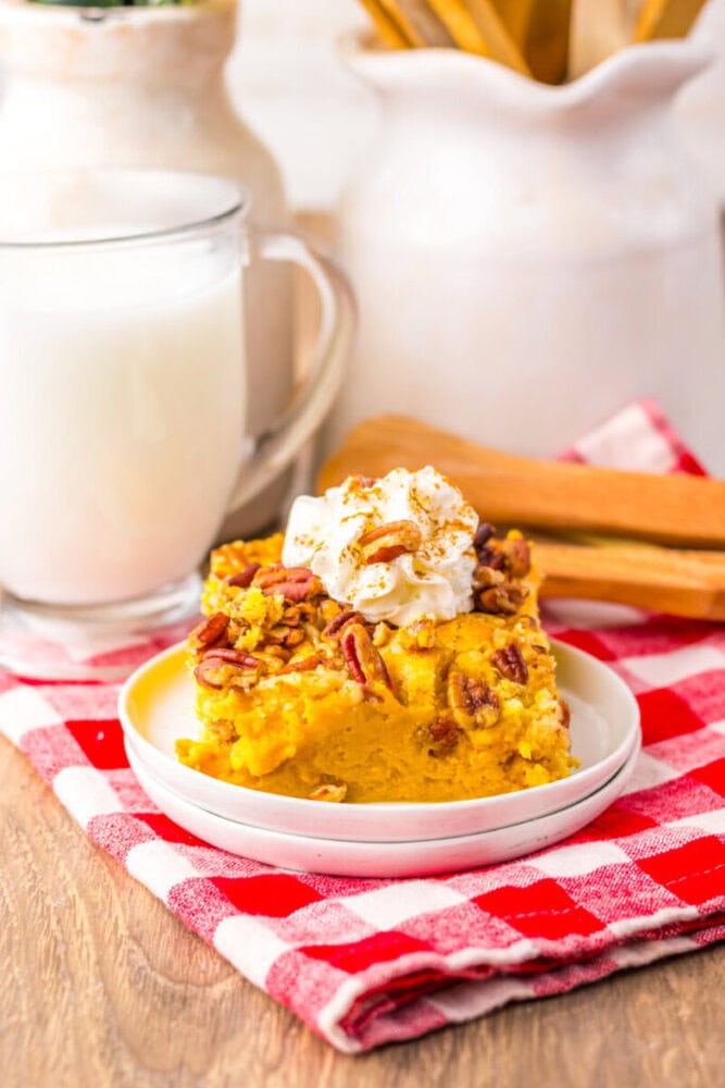 A slice of pumpkin dessert topped with whipped cream and pecans on a white plate, next to a glass of milk, set on a red and white checkered cloth.
