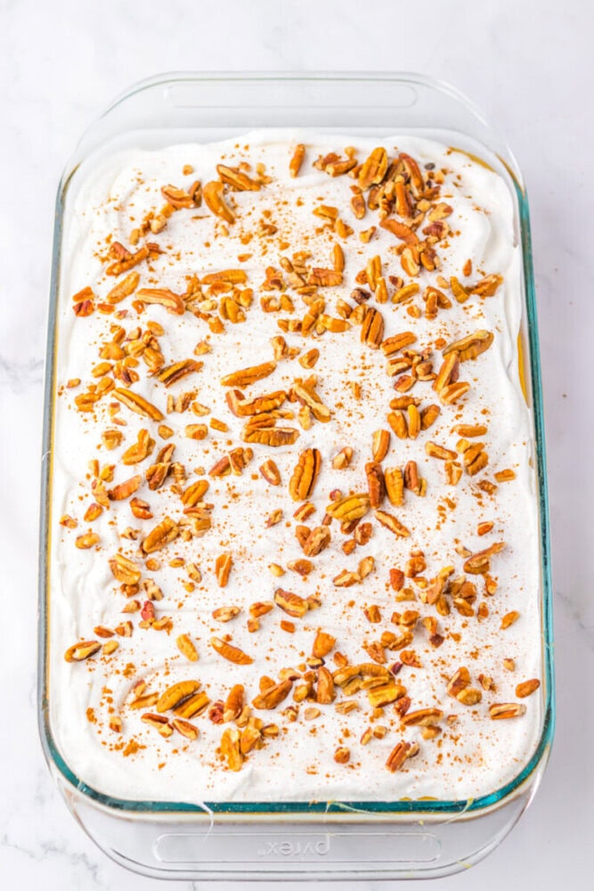 A glass baking dish filled with a dessert topped with whipped cream, chopped pecans, and a sprinkle of cinnamon on a white surface.